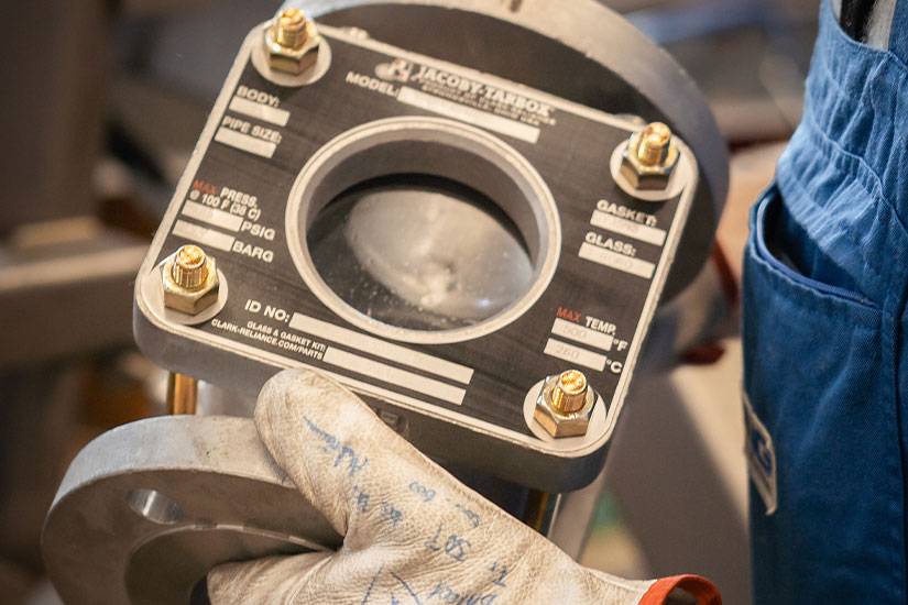 Ein Mann in blauen Overall und weißen Lederhandschuhen hält ein Druchflussanzeiger-Ventil von Jacoby Tarbox in den Händen.