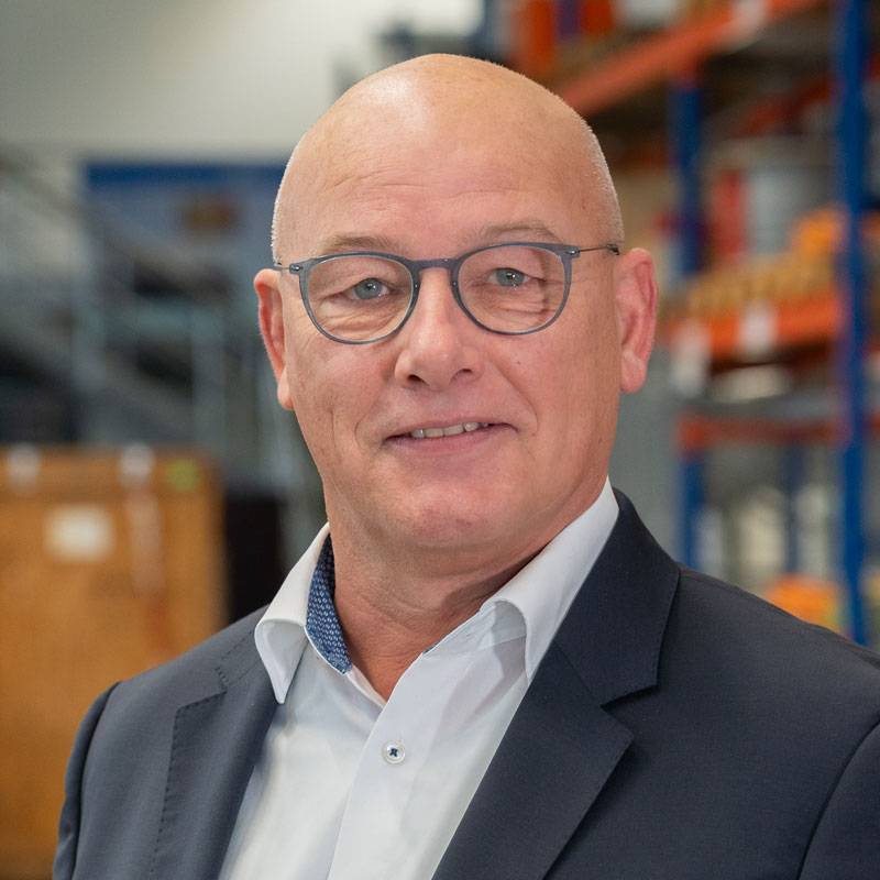 Zu sehen ist ein Mann in geschäftlicher Kleidung. Er trägt ein weißes Hemd, eine dunkle Krawatte und eine Brille mit dunklem Rahmen. Im Hintergrund befindet sich eine strukturierte graue Oberfläche. Der Mann lächelt freundlich in die Kamera.