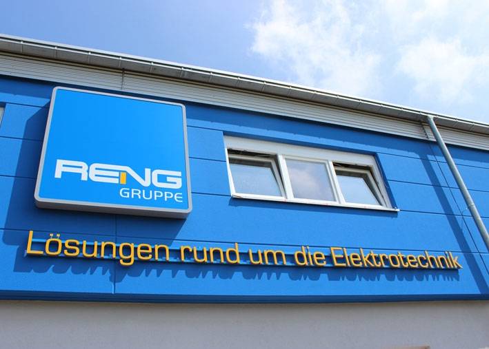 Ein blau vertäfeltes Gebäude mit weißen Fenstern. Auf der Vertäfelung ein blaues, quadratisches Schild mit abgerundeten Ecken und grauen Rand. Auf dem Schild in weißen Großbuchstaben geschrieben "Reng". Dabei ist der Aufschwung des Buchstabens N in Orange. Darunter ist klein rechts im Eck noch das Wort "Gruppe". Unter dem Schild ist noch eine Schrift auf die Wand montiert: "Lösungen rund um die Elektrotechnik".
