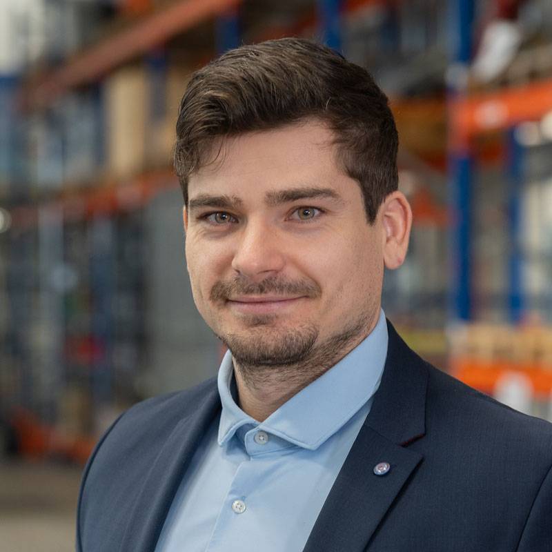 Studioaufnahme eines Mannes im hellblauen Hemd vor dunkelblauem Hintergrund. Er blickt leicht seitlich lächelnd in die Kamera. Das Bild vermittelt einen professionellen und modernen Eindruck.