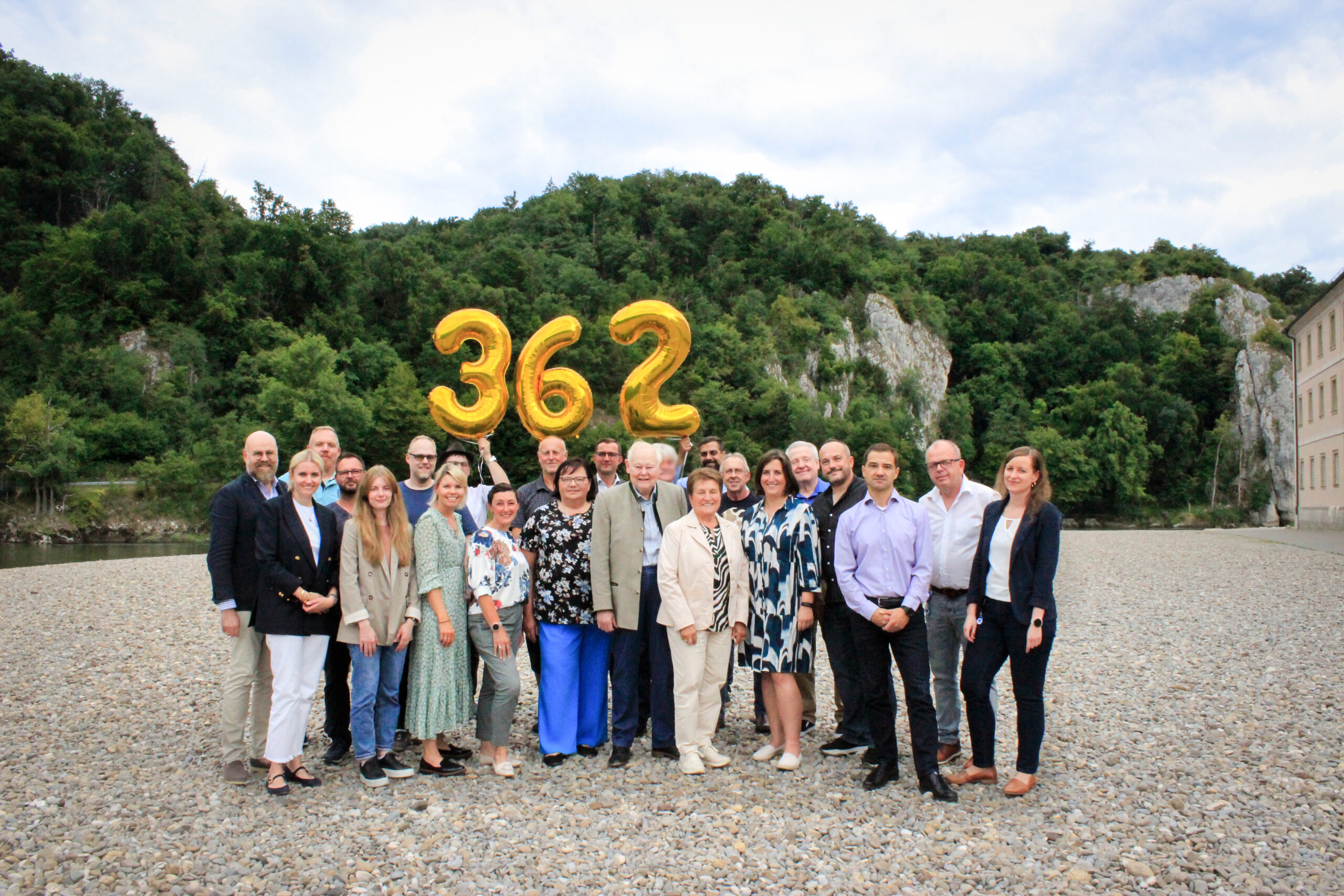 Zusehen ist eine Gruppe Menschen, die auf einer Kiesbank stehen. Die Personen in der hinteren Reihe halten goldene Zahlen als Luftballons hoch. Die Zahlen sind 3 6 2. Im Hintergrund befinden sich Felsen, Bäume und Büsche.