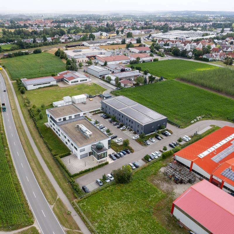 Luftaufnahme eines modernen Gewerbeareals mit mehreren Büro- und Produktionsgebäuden, großzügigen Parkflächen und umliegenden Feldern. Das Hauptgebäude besitzt einen markanten Eingangsbereich mit verglastem Turm. Die Umgebung zeigt weitere Gewerbehallen, Straßenanbindung und angrenzende Wohngebiete.