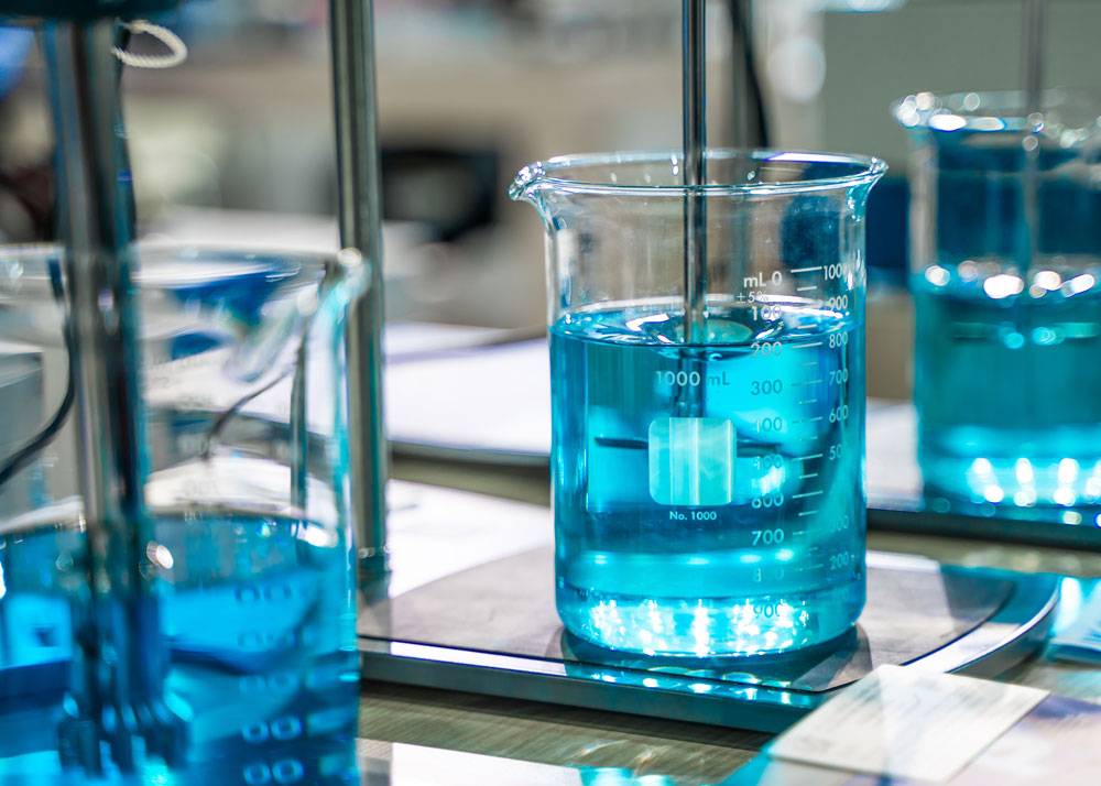 Das Bild zeigt ein großes Becherglas mit blauer Flüssigkeit, das auf einer Laborwaage oder einem Rührgerät steht. In der Flüssigkeit befindet sich ein Rührstab, der von oben in das Gefäß führt. Die Skala auf dem Glas ist gut sichtbar und zeigt ein Volumen von bis zu 1000 Millilitern. Im Hintergrund stehen weitere Bechergläser mit ähnlicher Flüssigkeit. Die Szene vermittelt einen typischen Arbeitsschritt in einem chemischen oder analytischen Labor.