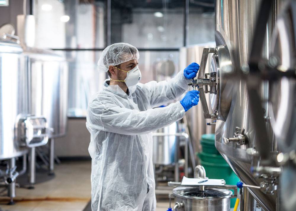 Ein Mitarbeiter in Schutzkleidung – bestehend aus Overall, Haube, Maske und blauen Schutzhandschuhen – öffnet die Revisionsöffnung eines großen Edelstahltanks in einer Produktionshalle. Im Hintergrund stehen weitere glänzende Tanks und technische Anlagen. Der Mitarbeiter arbeitet konzentriert an der Armatur, während Werkzeug und ein Behälter für Probenahmen bereitstehen.