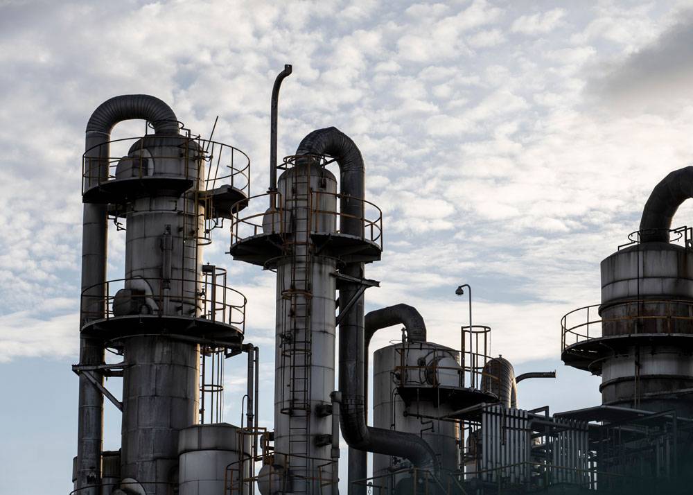 Das Bild zeigt mehrere hohe, zylindrische Industrieanlagen mit angeschlossenen Rohrleitungen, Plattformen und Sicherheitsgeländern. Die Metallkonstruktionen sind witterungsgeprägt und über Leitern sowie Wartungsbühnen zugänglich. Dicke Leitungsbögen und Abzüge verlaufen zwischen den Türmen. Der Himmel im Hintergrund ist bewölkt und sorgt für eine kontrastreiche Silhouette der Anlage.