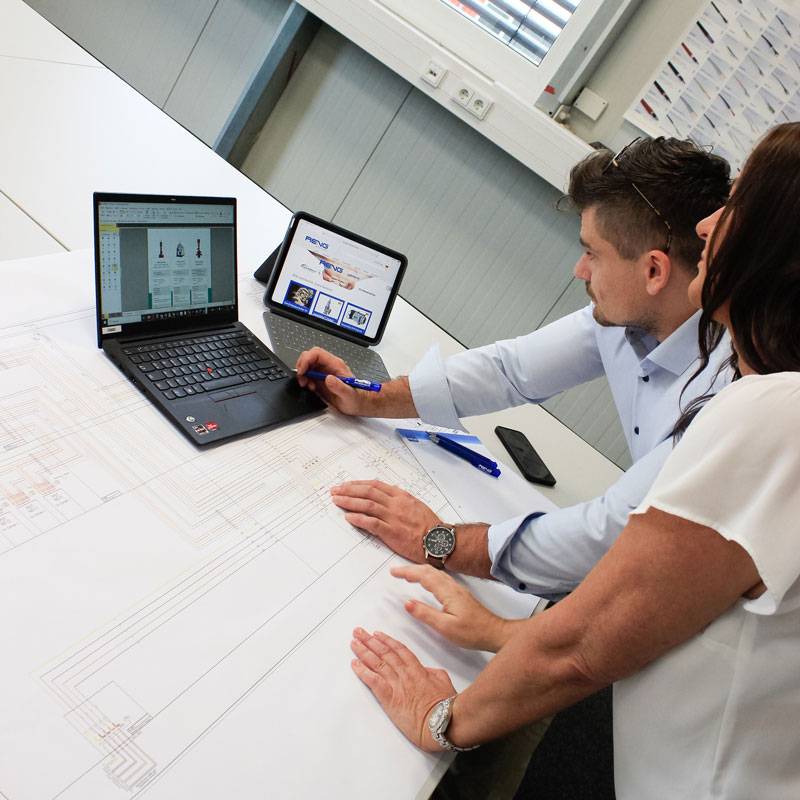 Ein Mann und eine Frau sind über einen großen technischen Plan und einem Laptop gebeugt und und sehen sich Veniltdaten auf dem Bildschirm gemeinsam an.