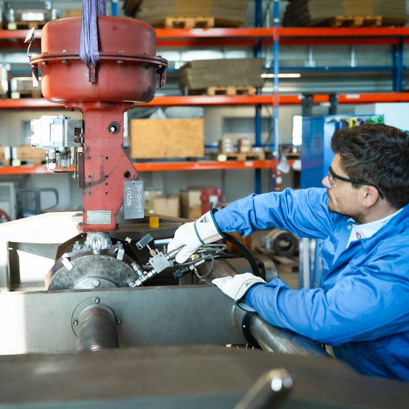Ein Mann im blauen Overall, weißen Lederhanschuhen und transparenter Sicherheitsbrille moniert an einem Masoneilan Ventil Schläuche.