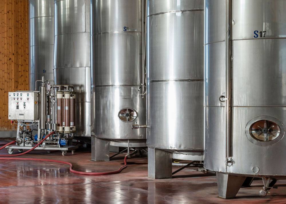 Das Bild zeigt mehrere große Edelstahltanks in einer Produktions- oder Lagerhalle. Die Tanks stehen auf stabilen Stahlfüßen und verfügen über runde Revisionsöffnungen sowie seitliche Anschlüsse. Links im Bild befindet sich eine Technik- und Filtereinheit mit Schläuchen und einer Steuerkonsole. Der Boden ist glatt und leicht reflektierend, während die Umgebung aus Holzverkleidung und industriellen Elementen besteht. Die Szene vermittelt typische Abläufe in der Lebensmittel-, Getränke- oder Chemieproduktion.