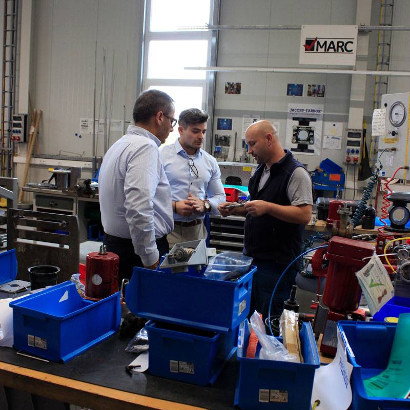 3 Männer stehen sich gegenüber in einer Werkstatt, in der viel Chaos herrscht und besprechen etwas. Die Gesichter der Männer sind ernst.