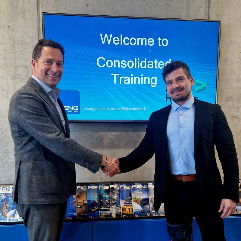 Zwei Männer in Anzug stehen vor einem großen Bildschirm und einem Zeitschriftenregal mit Reng Magazinen und reichen sind die Hand. Die Männer lächeln beide in die Kamera. Auf dem Bildschirm ist ein Bild mit blauem Hintergrund und Schrift "Welcome to Consolidated Training" sowie das Logo der Reng Gruppe und Baker Hughes.