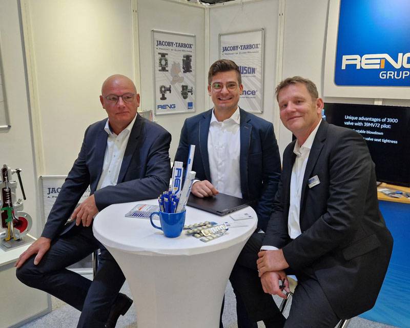 Drei Männer in schwarzen Anzügen sitzen auf Barhockern hinter einem Stehtisch auf einem Messestand der Reng Gruppe. Der linke Mann blickt ernst in die Kamera, während der mittere Mann in die Kamera grinst und der rechte nur schmunzelt.