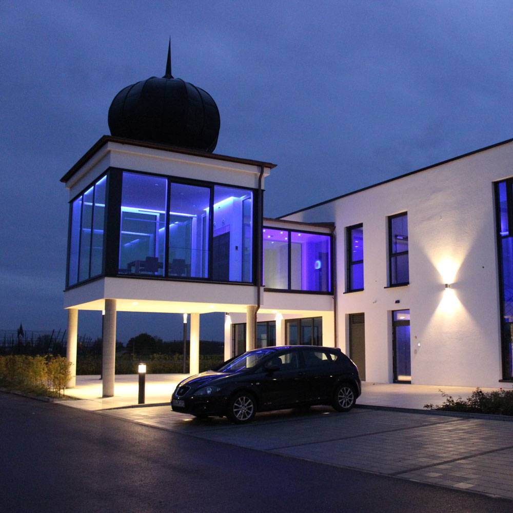 Das Bild zeigt ein modernes Firmengebäude in der Abenddämmerung. Die Architektur verfügt über einen verglasten oberen Bereich mit intensiver blauer Innenbeleuchtung, der von Säulen getragen wird. Im Vordergrund steht ein dunkles Auto auf dem Parkplatz. Außenbeleuchtung und klare Linien unterstreichen den modernen Charakter des Gebäudes.