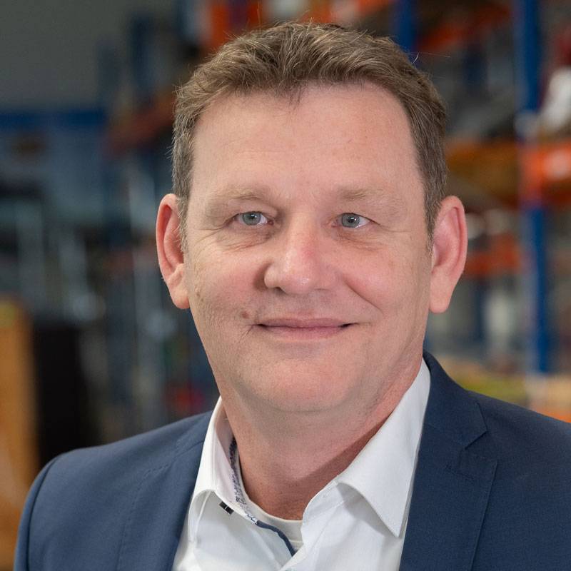 Professionelles Portrait eines Mannes in weißem Hemd und dunkelblauer Krawatte vor einer grauen Wand. Er steht leicht seitlich zur Kamera und lächelt freundlich.